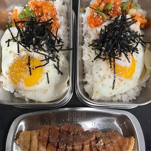 Two Korean BBQ rice bowls with fried eggs, kimchi, seaweed, and a side of sliced pork belly.