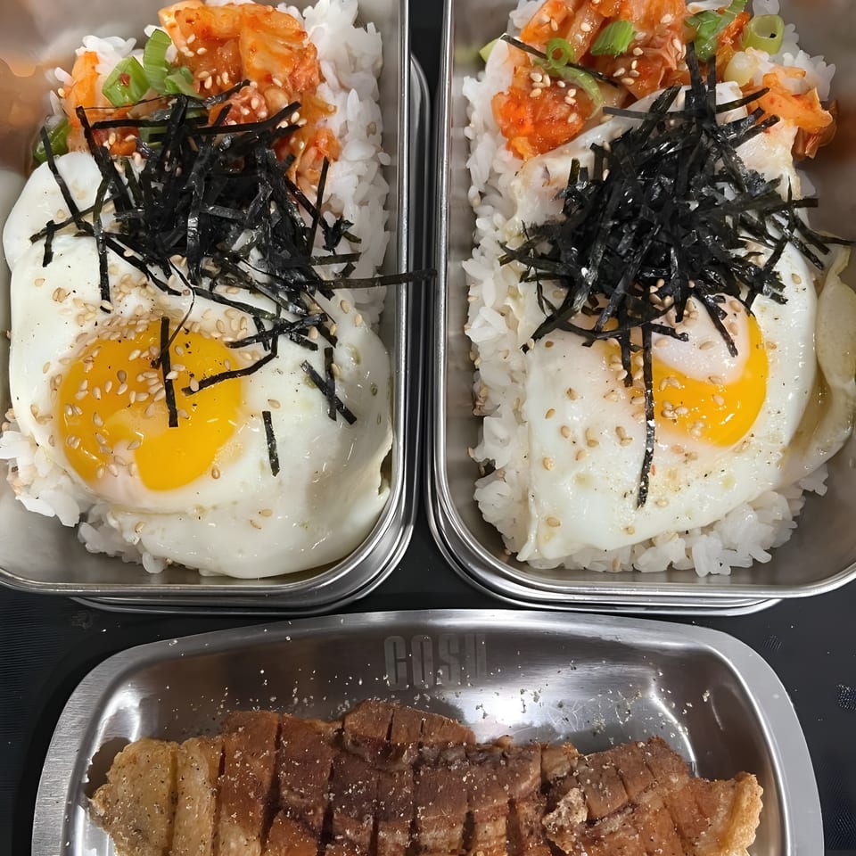 Two Korean BBQ rice bowls with fried eggs, kimchi, seaweed, and a side of sliced pork belly.