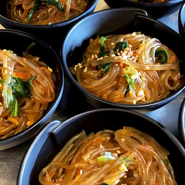 Multiple small black bowls filled with Korean japchae noodles, garnished with sesame seeds and greens.